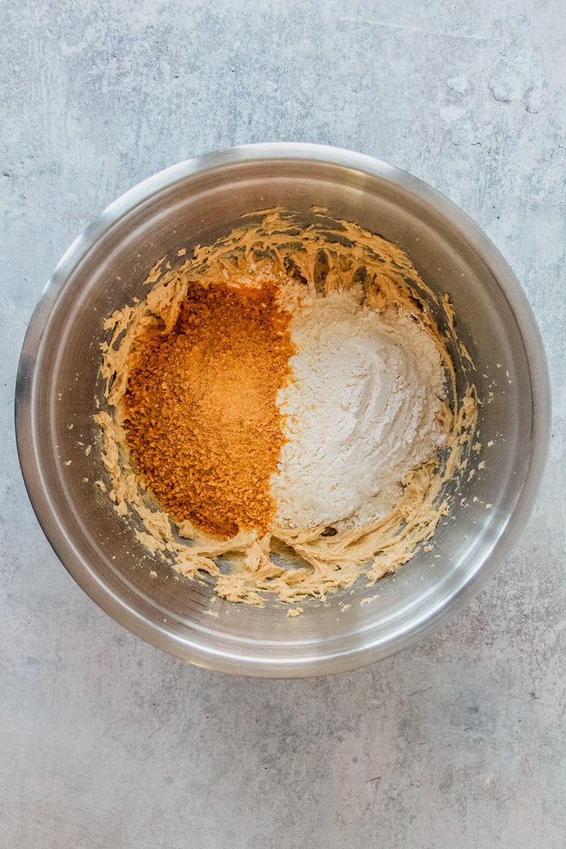 Crushed toasted cornflakes and dry ingredients sit on top of whipped cookie dough in a stainless steel bowl on a gray surface.