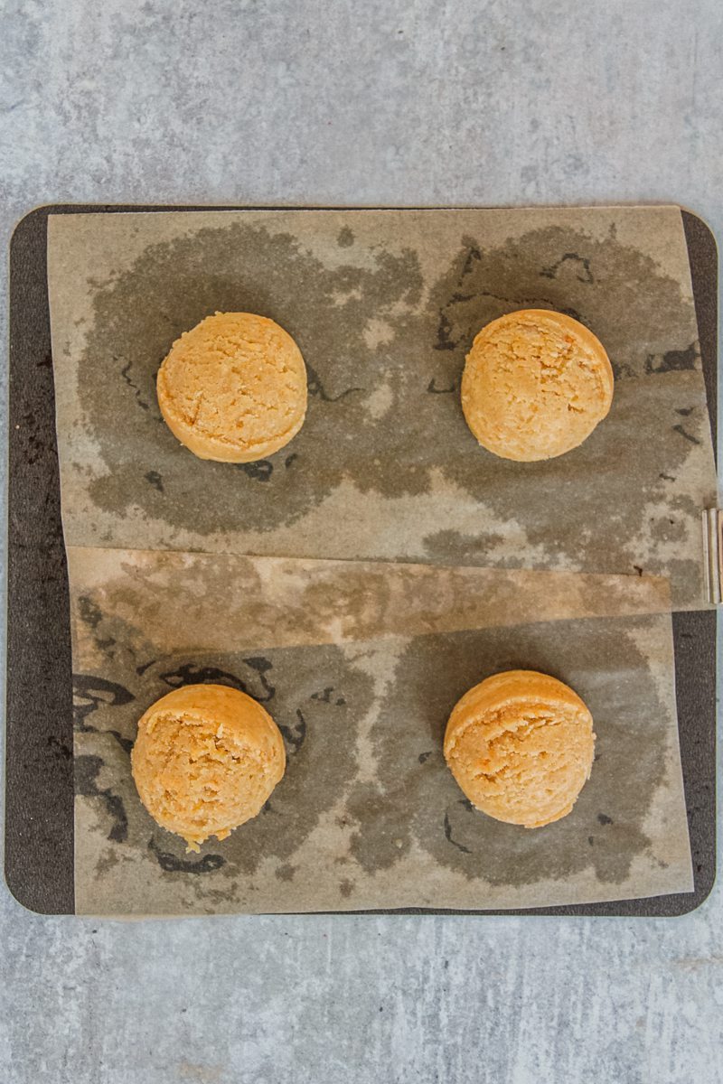 Unbaked cornflake cookie dough balls sit on a lined square metal baking tray on a gray surface.