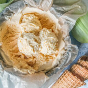 Scoops of no churn corn ice cream sit on top of a lined round metal tin with cones and corn cobs beside on a light gray surface.