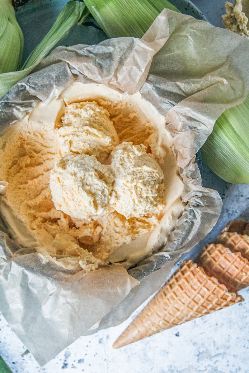 Scoops of no churn corn ice cream sit on top of a lined round metal tin with cones and corn cobs beside on a light gray surface.