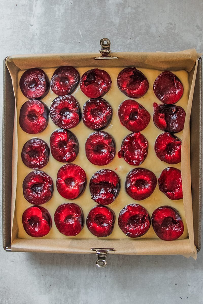 Cake batter with halved plums sit cut side up pressed into the batter in parchment paper in a square metal tin on a gray surface.