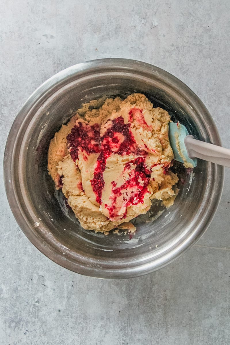 Homemade raspberry jam sits on top of cookie dough in a stainless steel bowl on a gray surface.