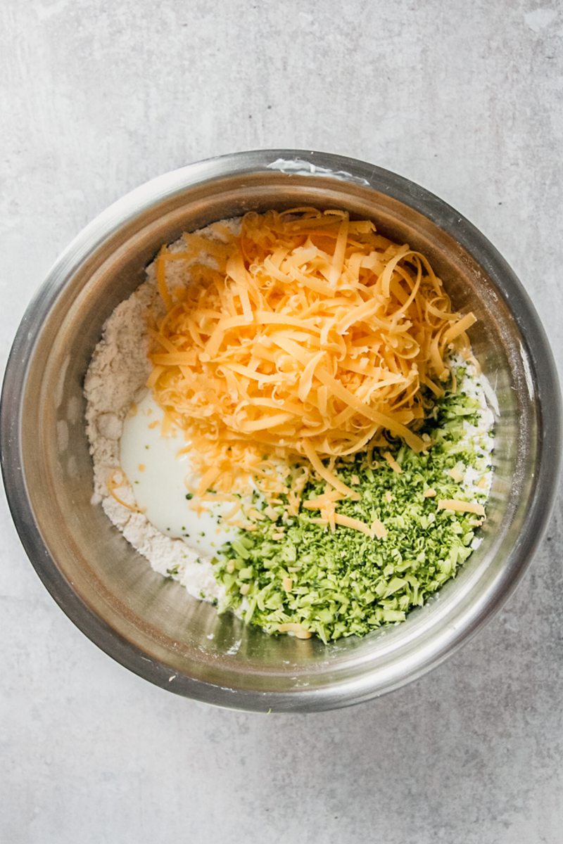 Grated cheddar cheese and finely chopped broccoli sit top of a butter and flour crumb mixture in a stainless steel bowl on a gray surface.