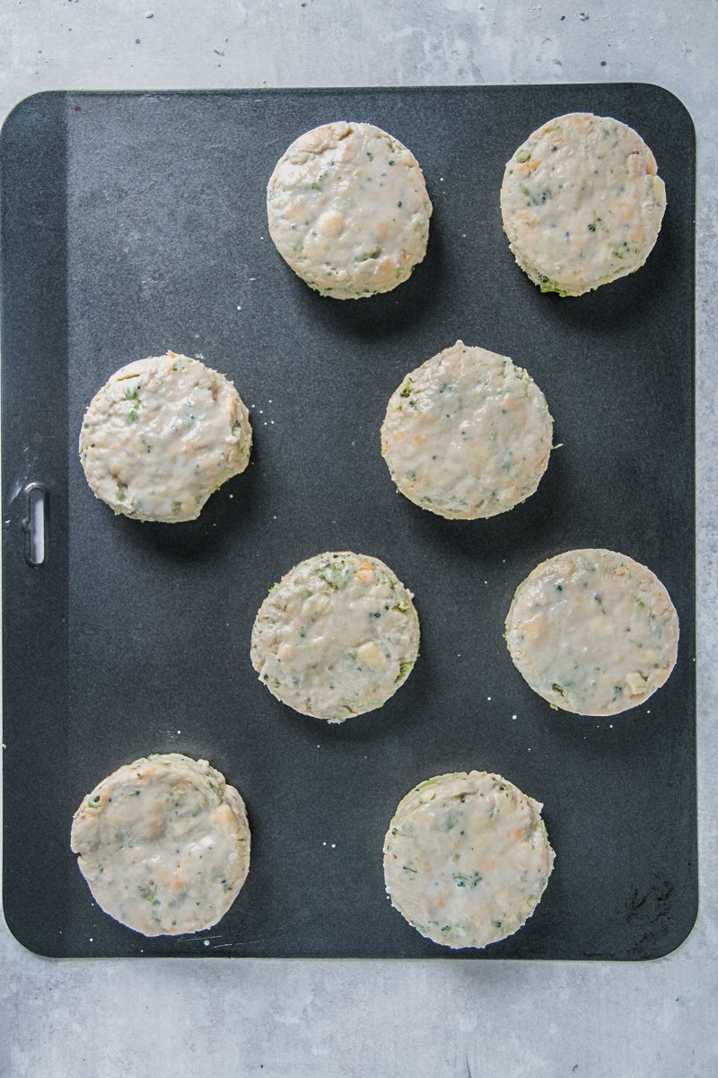 Round cheddar biscuits sit on a baking tray on a gray surface.