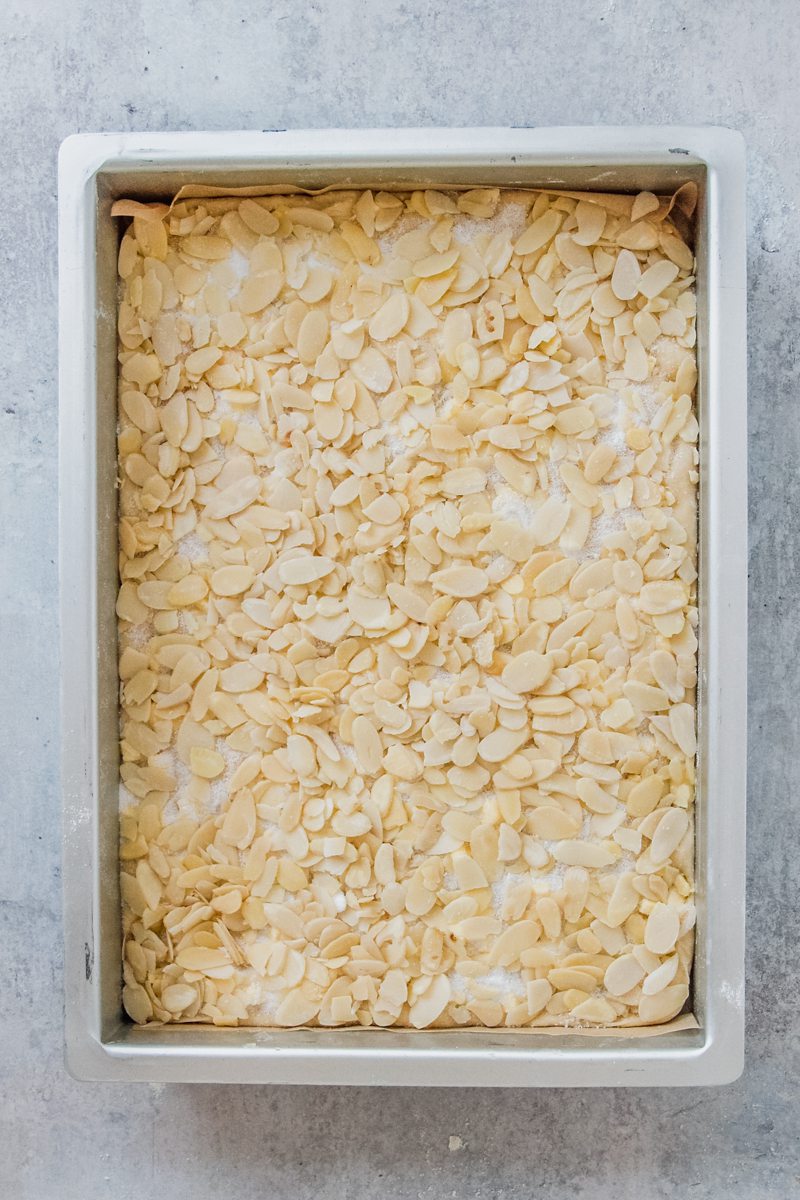 Sliced almonds sit on top of sugar on a yeast cake in a rectangle metal baking tin on a gray surface.