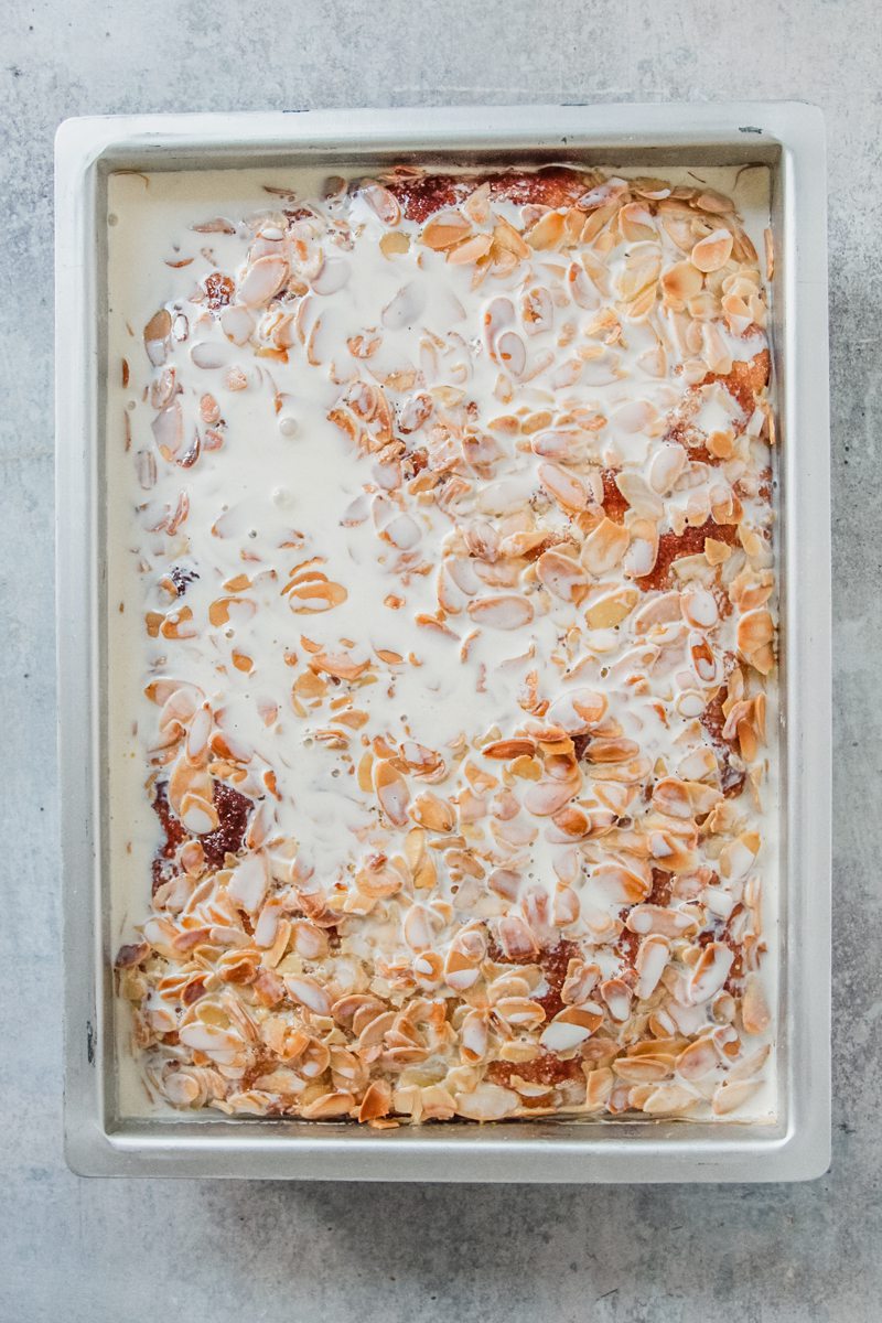 Cream sits on top of a grolen baked German butter cake in a rectangle metal baking tin on a gray surface.