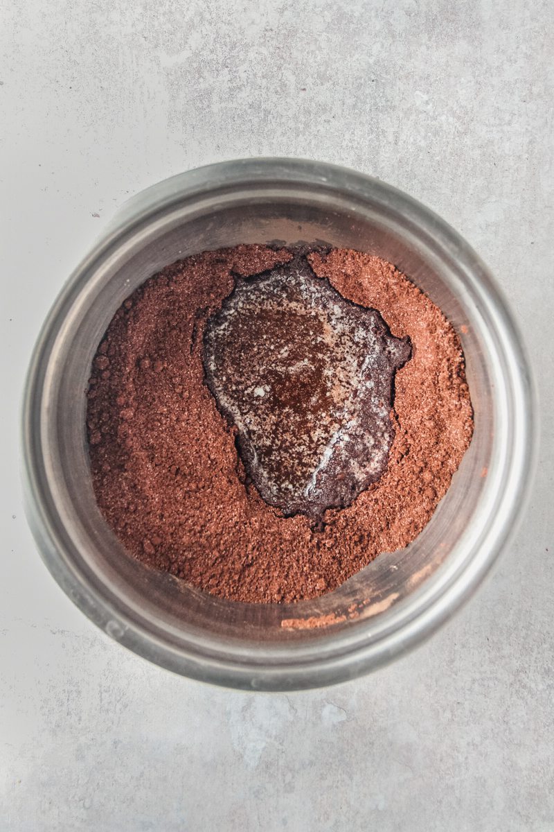 A crushed graham cracker cocoa mixture and melted butter sits in a stainless steel bowl on a gray surface.