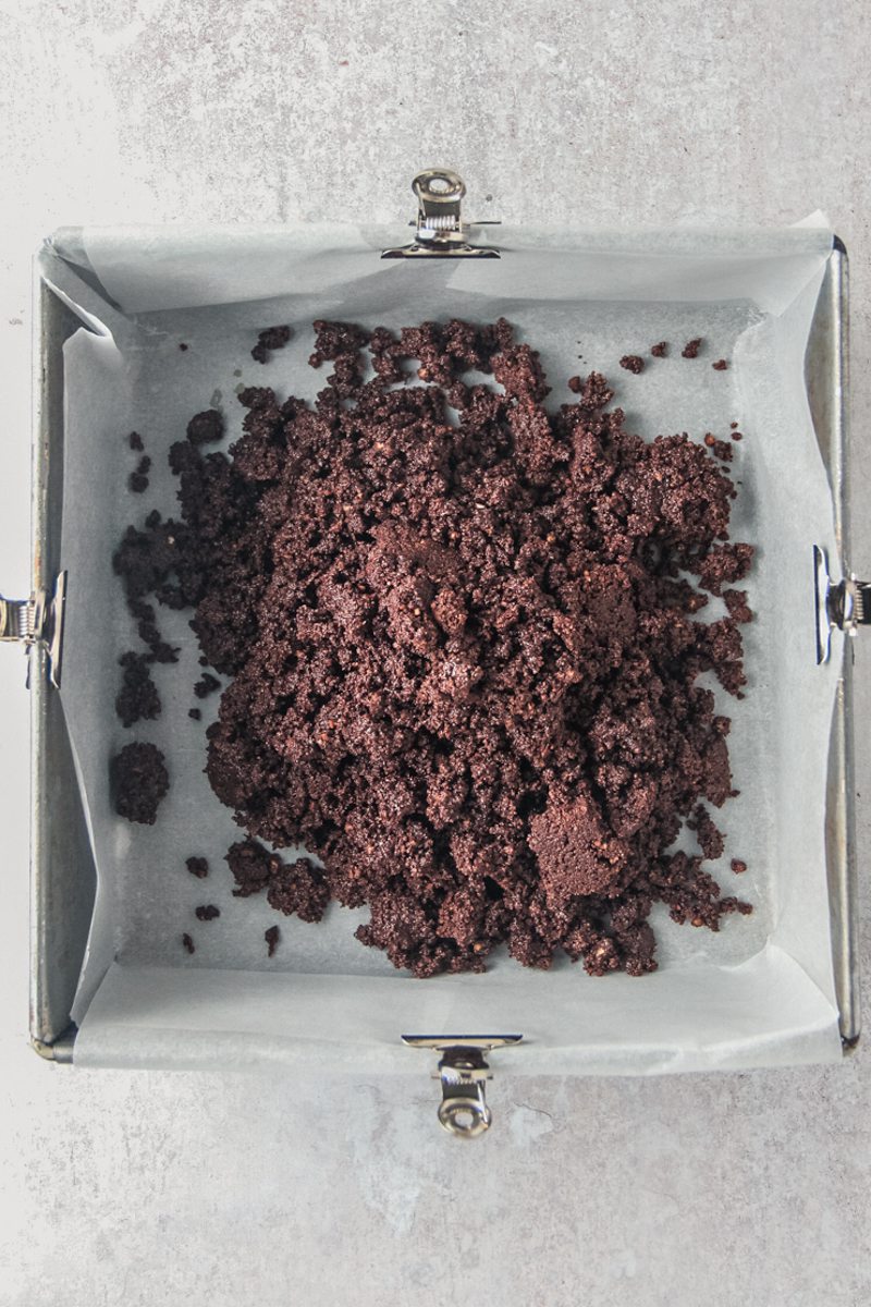 A graham cracker cocoa mixture sits in a rough pile in a lined square metal baking tin on a gray surface.