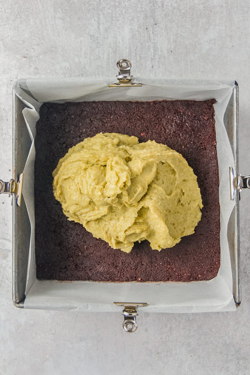 A pistachio buttercream sits in the center of a pressed crushed graham cracker cocoa base in a lined square metal baking tin.