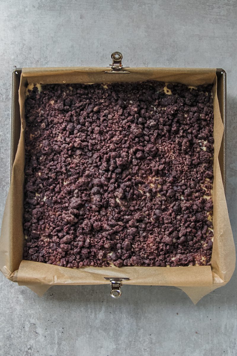 Blueberry streusel sits on top of cake batter in a lined square metal baking tin on a gray surface.