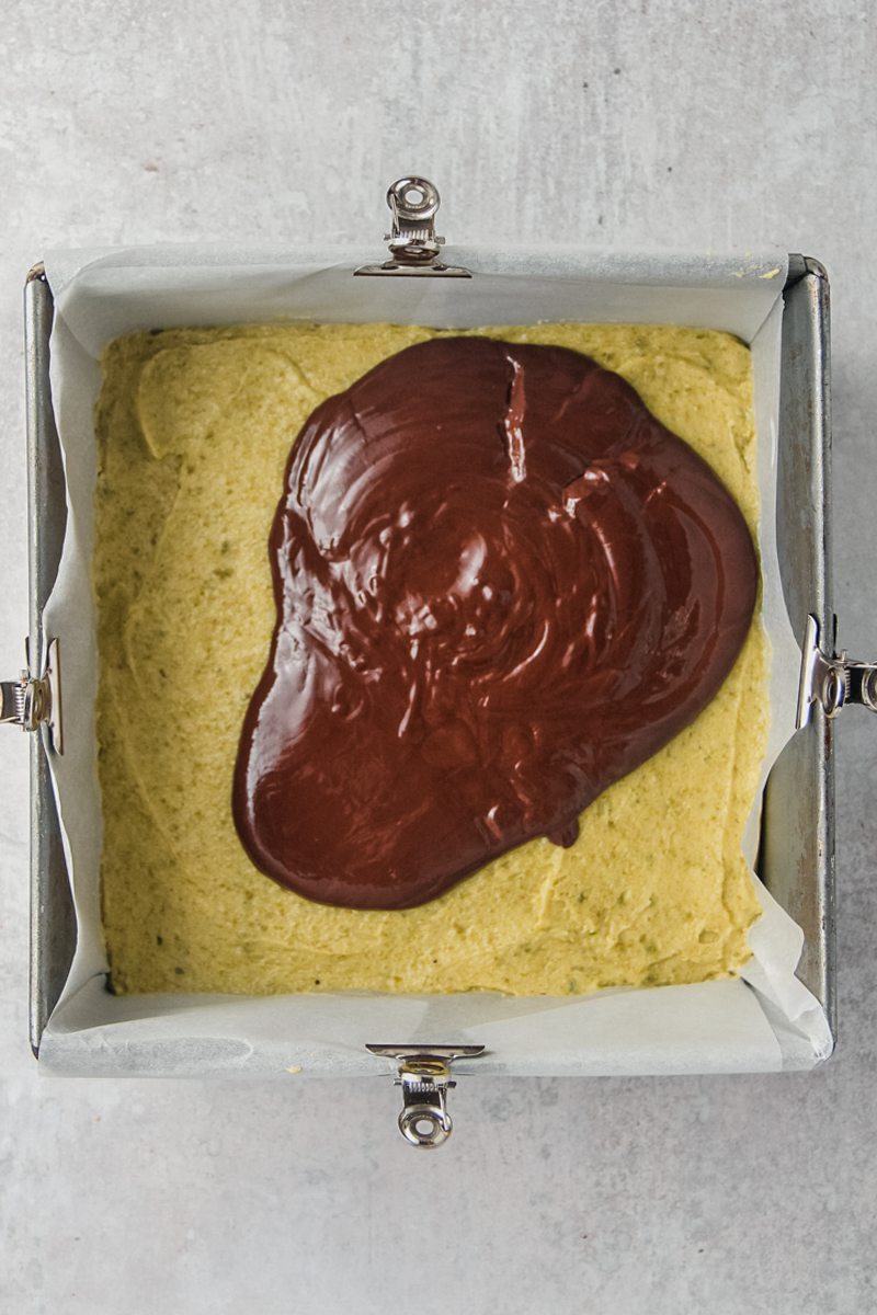 Melted chocolate sits on top of a smooth layer of pistachio buttercream in a lined square metal baking tin on a gray surface.