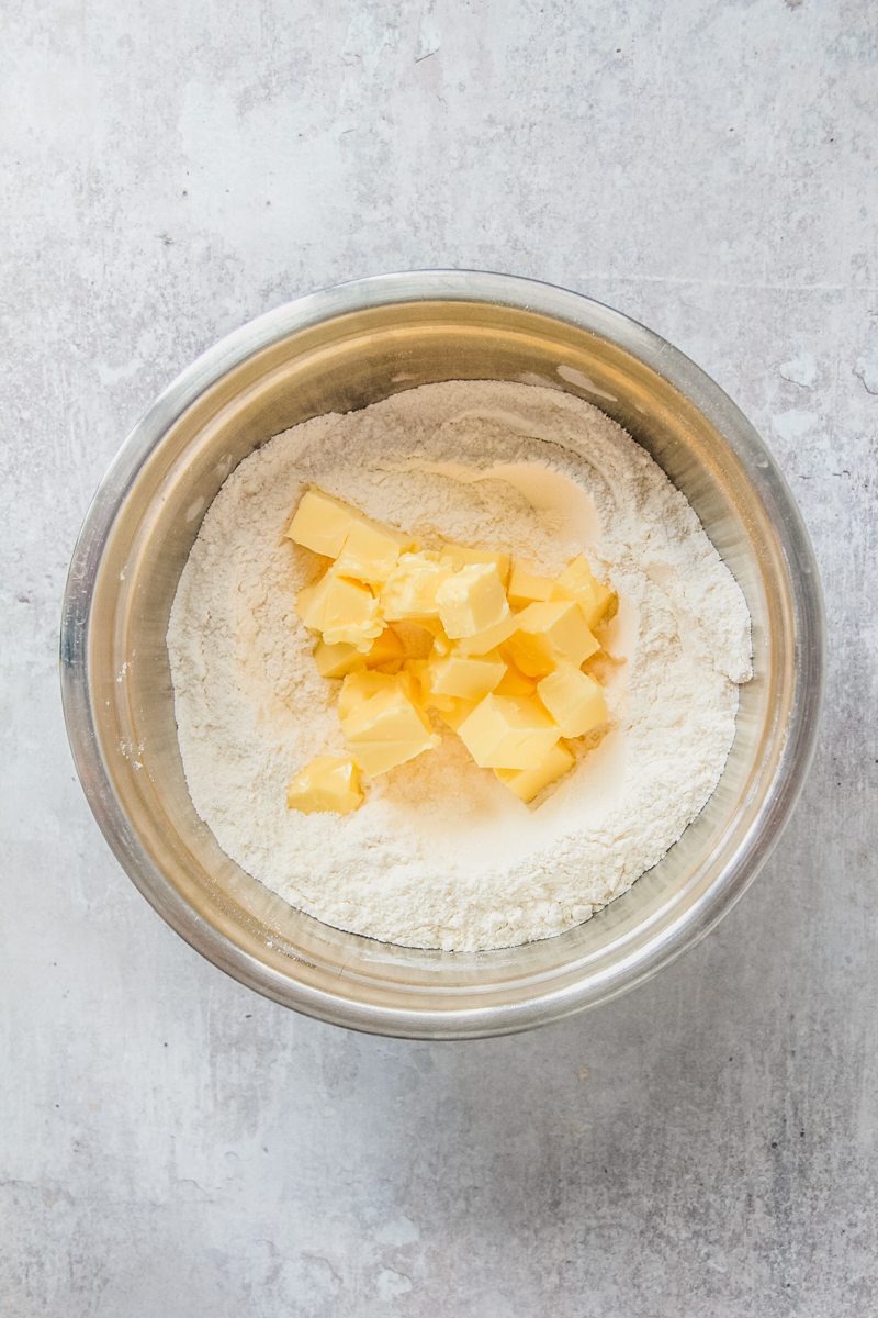 Cubes of butter sit in the center of a dry mixture in a stainless steel bowl on a gray surface.