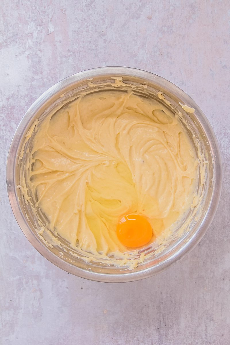 An egg sits on top of a vanilla cake batter in a stainless steel bowl on a gray surface.