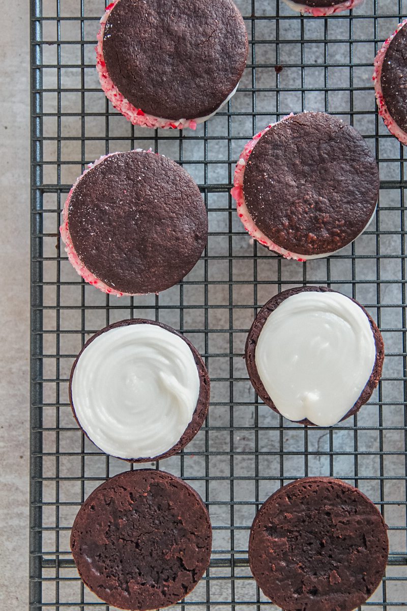 Peppermint icing coats the top of two cookies with other sandwiched cookies and the edges coated in crushed candy canes on a cooling rack.