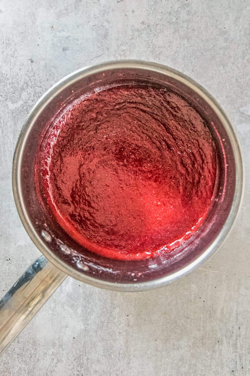 A reduced strawberry mixture sits in a stainless steel saucepan on a gray surface.