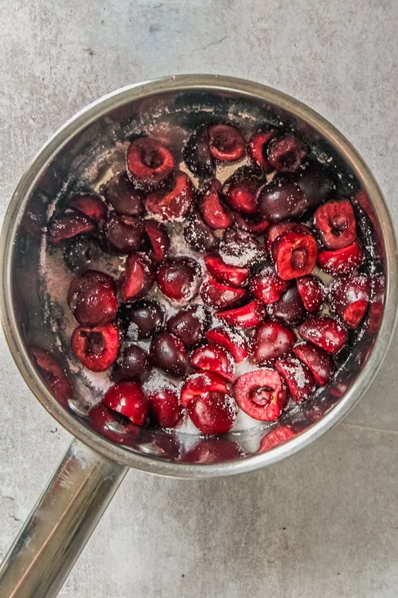 Halved cherries sit in a stainless steel saucepan coated in sugar on a gray surface.