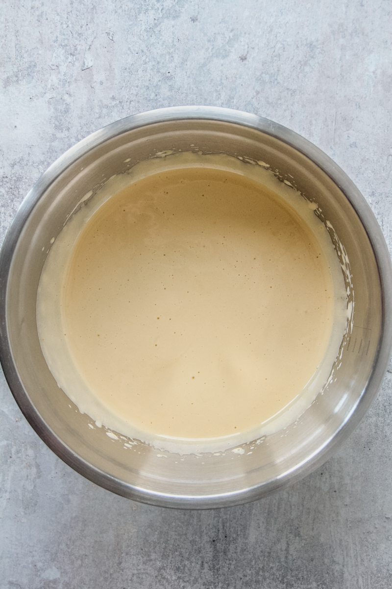 Pale whipped eggs and sugar sit in a stainless steel bowl on a gray surface.