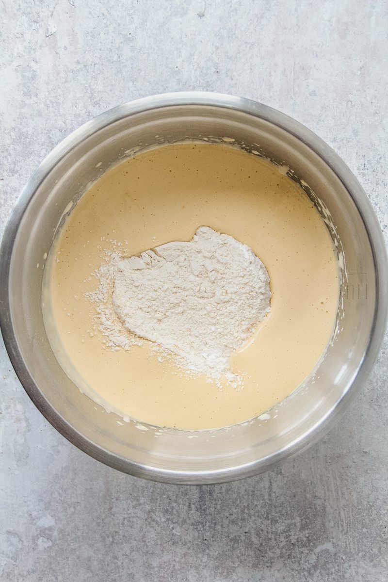 Dry ingredients sit in the center of a whipped egg and olive oil mixture in a stainless steel bowl on a gray surface.