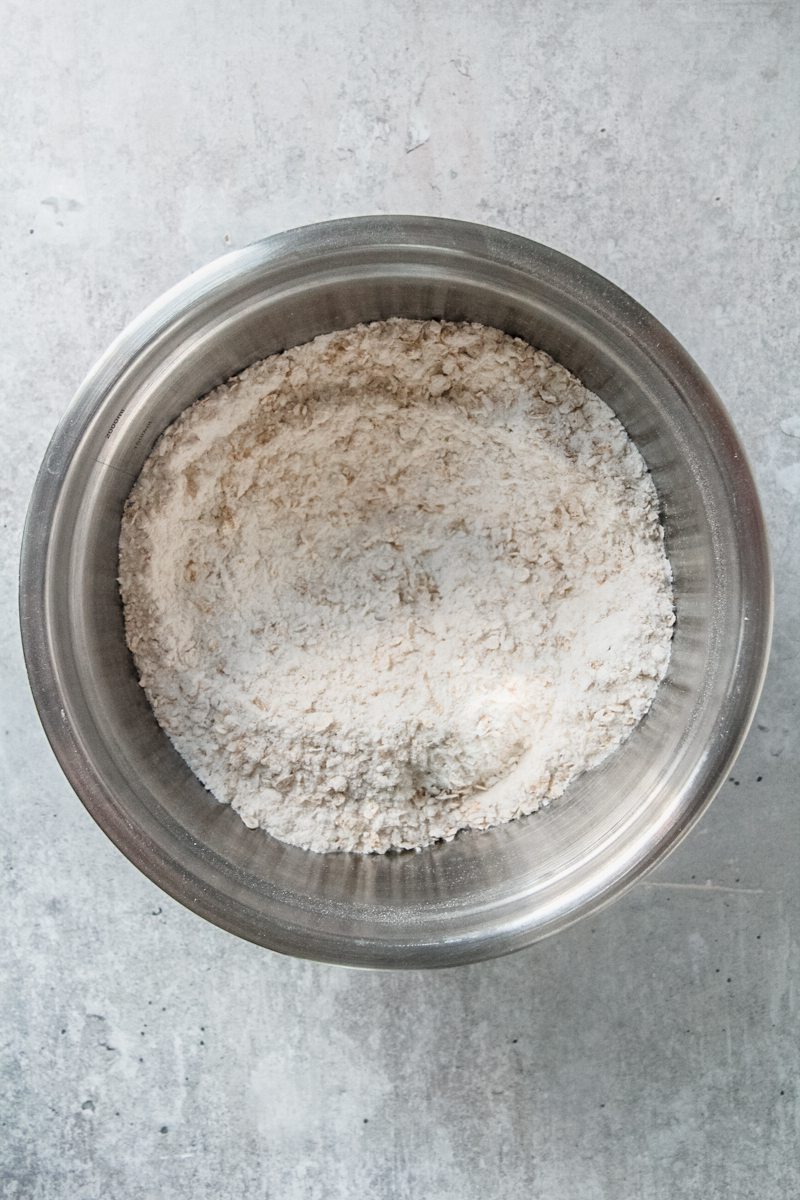 Flour and oats sit stirred with a well in the center of a stainless steel bowl on a gray surface.