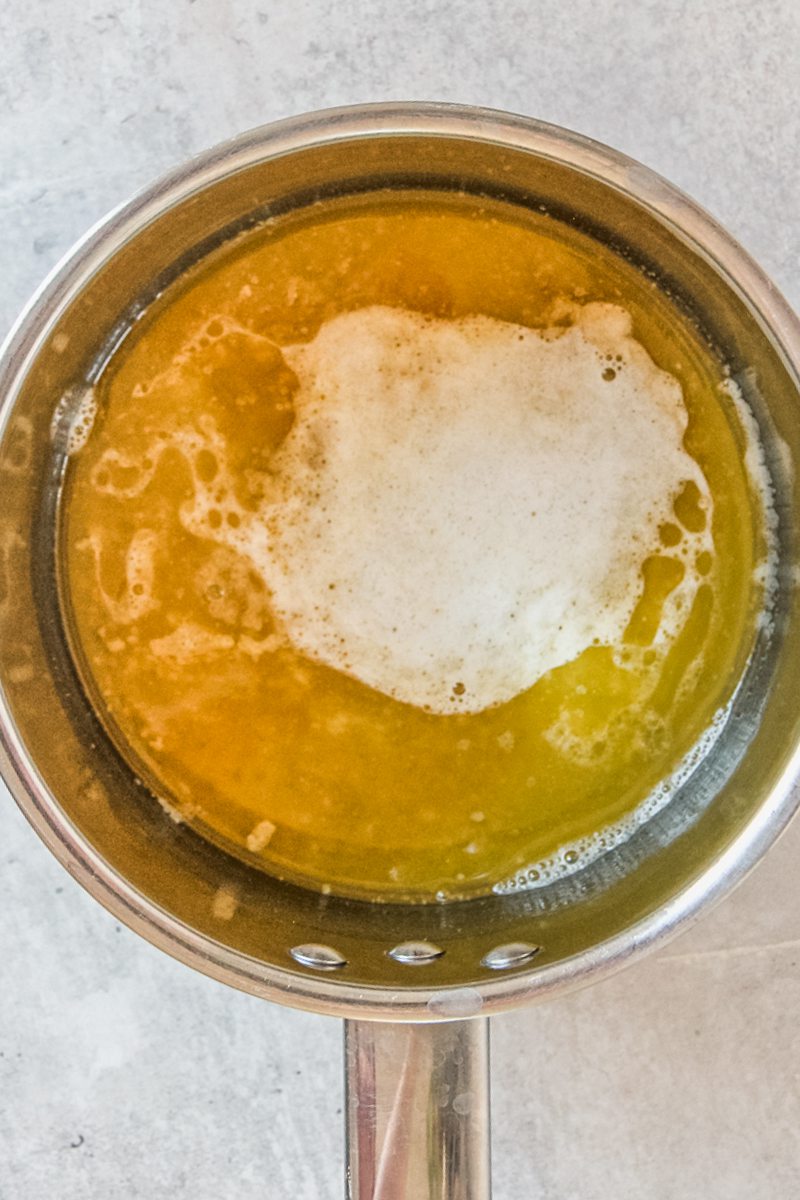 Melted butter has baking soda bubbles in the center in a stainless steel saucepan on a gray surface.