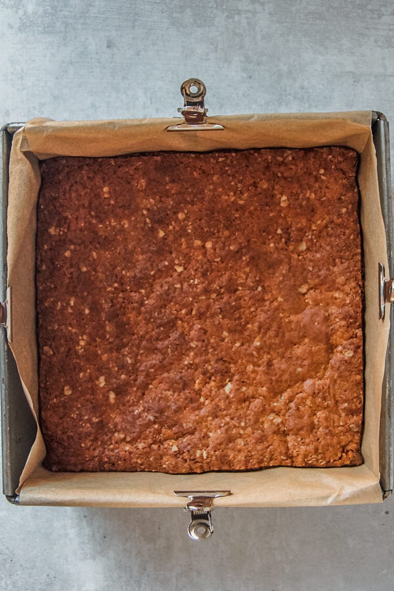 Golden baked anzac biscuit bars sit in a lined square metal baking tin on a gray surface.