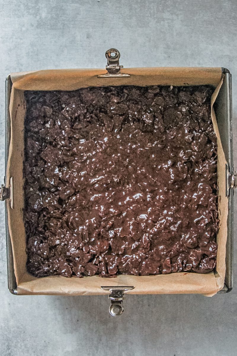 A cornflake chocolate mixture sits on top of anzac biscuit bars in a lined square metal baking tin on a gray surface.