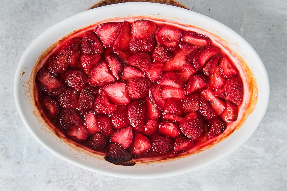 An oval baking dish sits on a light gray surface with deep red roasted strawberries inside.