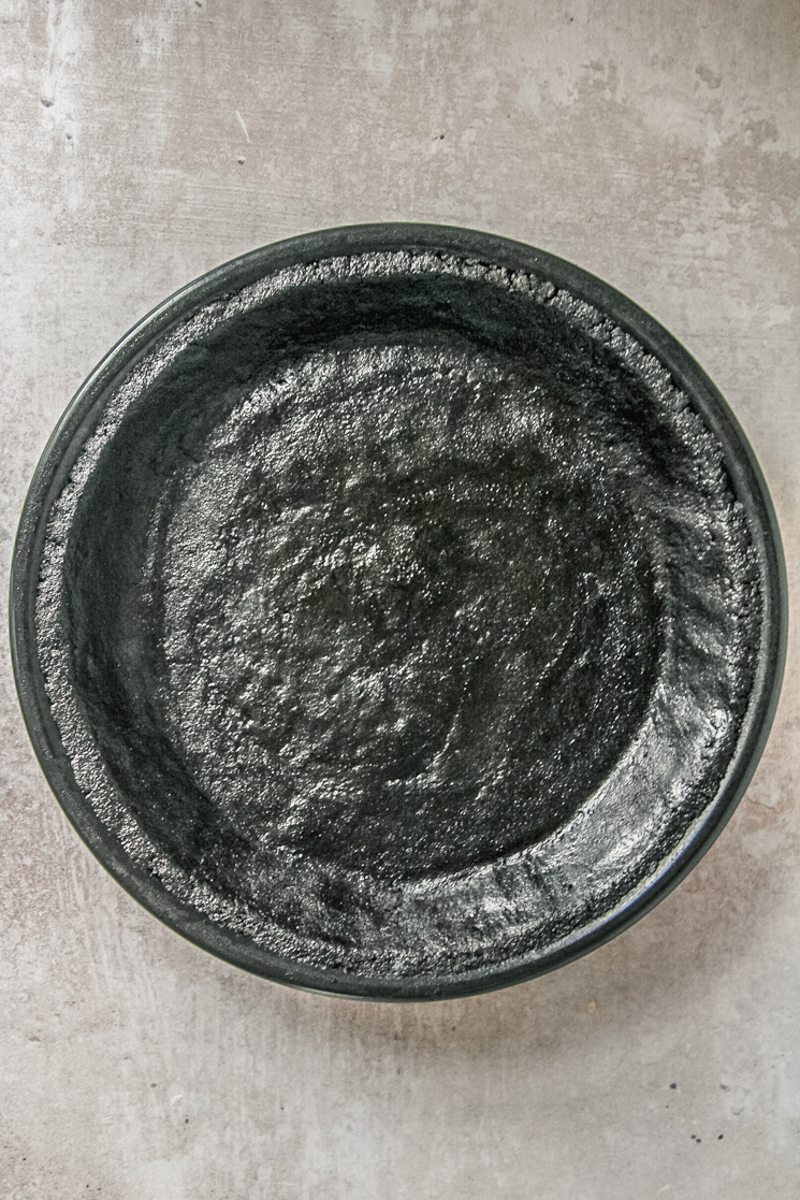 A oreo pie crust sits pressed into a black metal pie dish on a gray surface.