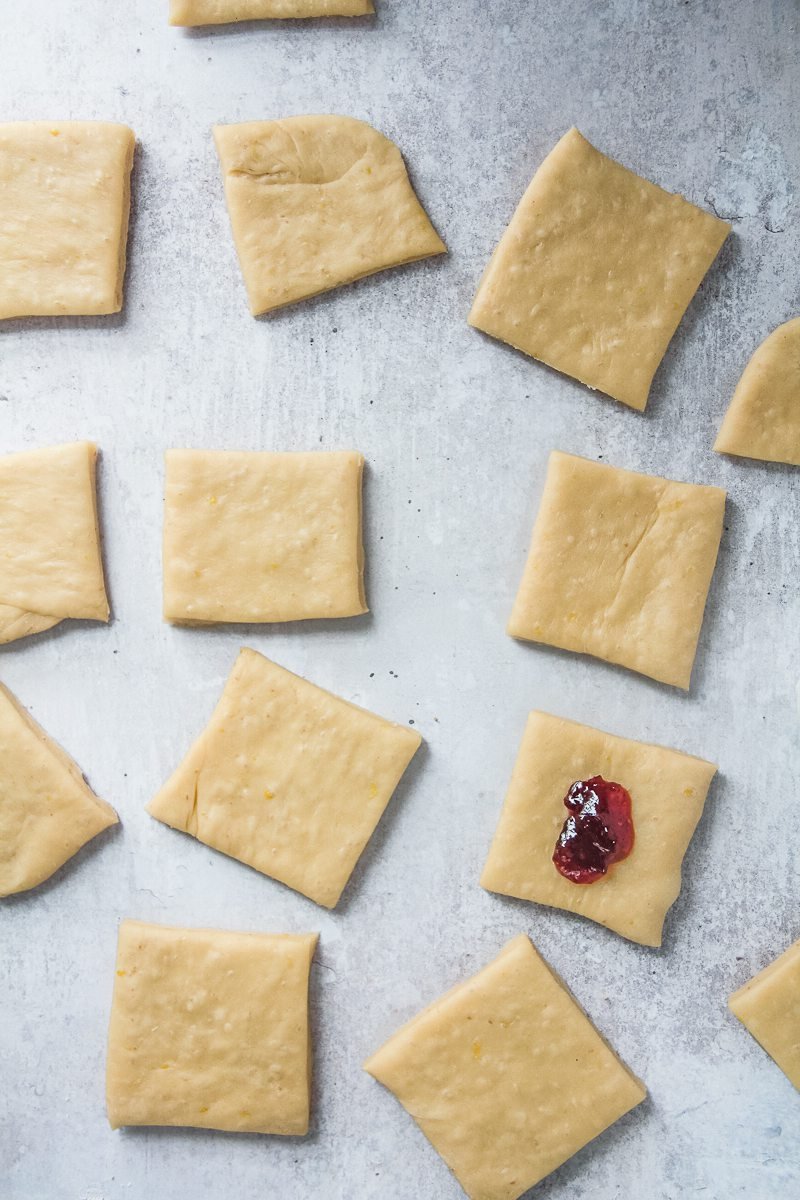 Squares of dough sit on a gray surface with one square with a dollop of jam in the center.