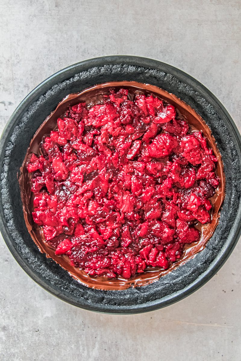 Cooked roughly chopped cherries sit on top of a chocolate pudding in a oreo crust sitting in a black metal pie dish on a gray surface.