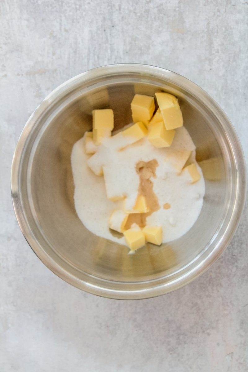 Cubes of butter, sugar and vanilla extract sit in a stainless steel bowl on a gray surface.