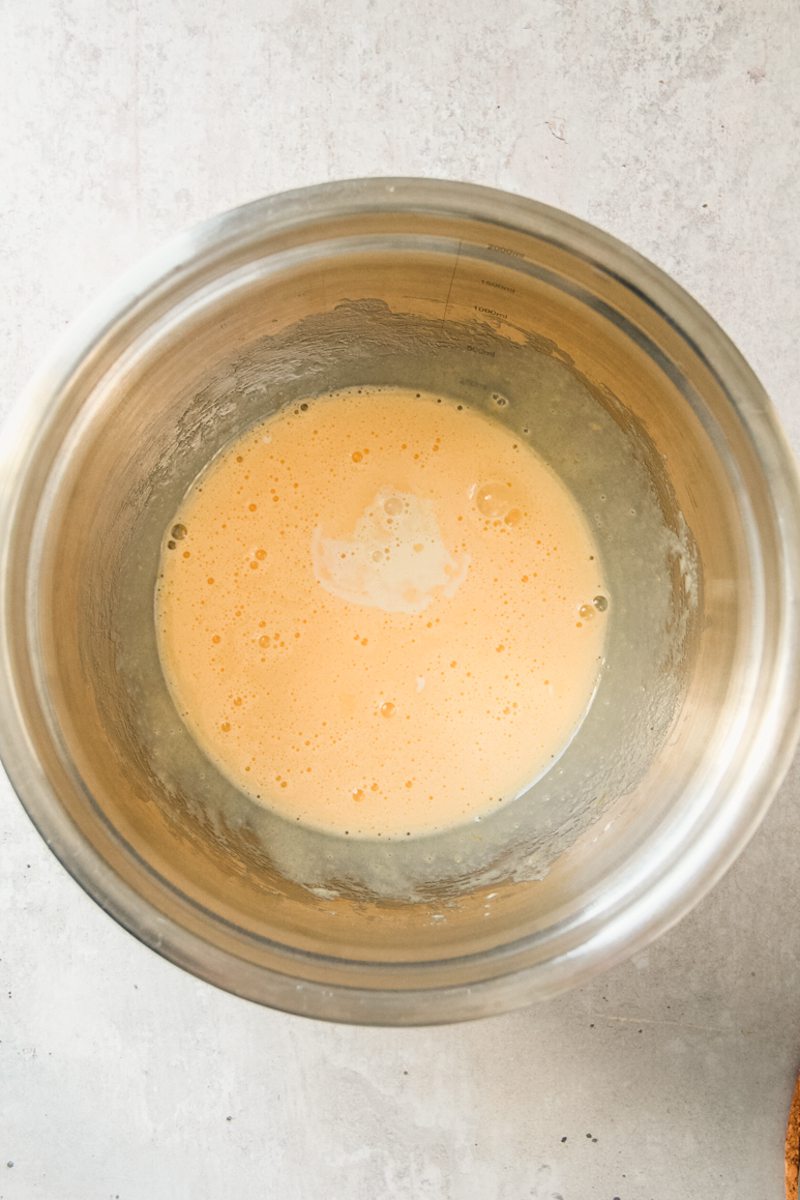 Whipped egg yolks and sugar sit in a stainless steel bowl on a gray surface with a little warm cream in the center.