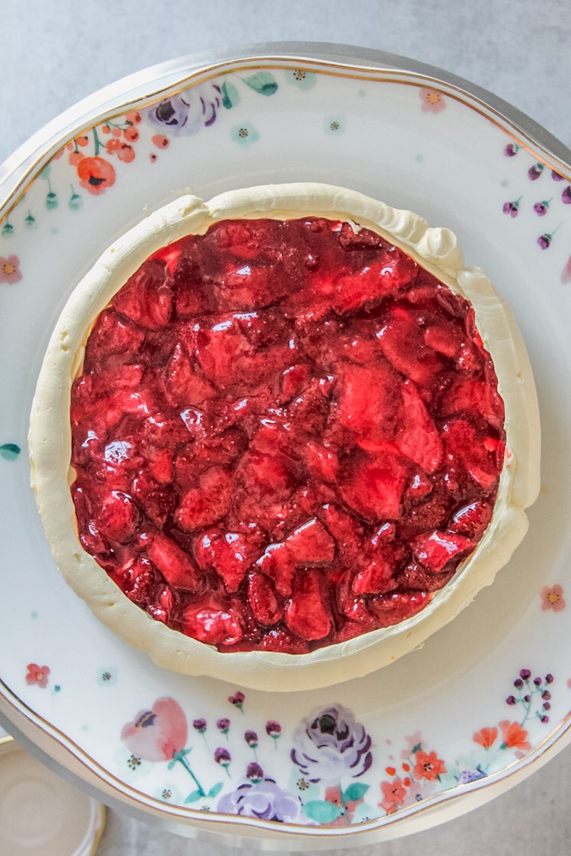 Roasted strawberries sit on the top of a cake with a Swiss meringue buttercream border on a floral ceramic plate above a gray surface.