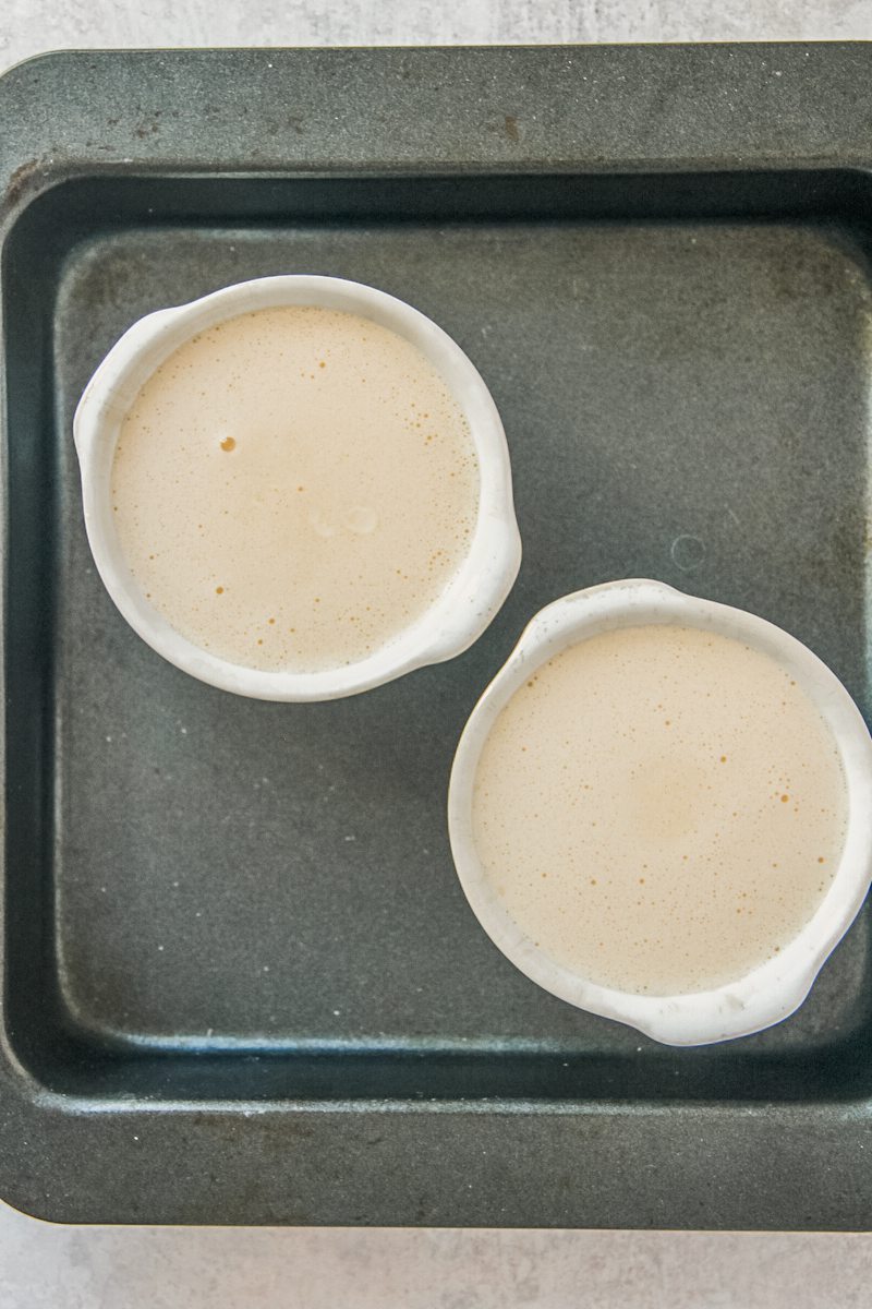 Earl gray creme brulee sits in individual ramekins in a square baking tin before baking on a gray surface.