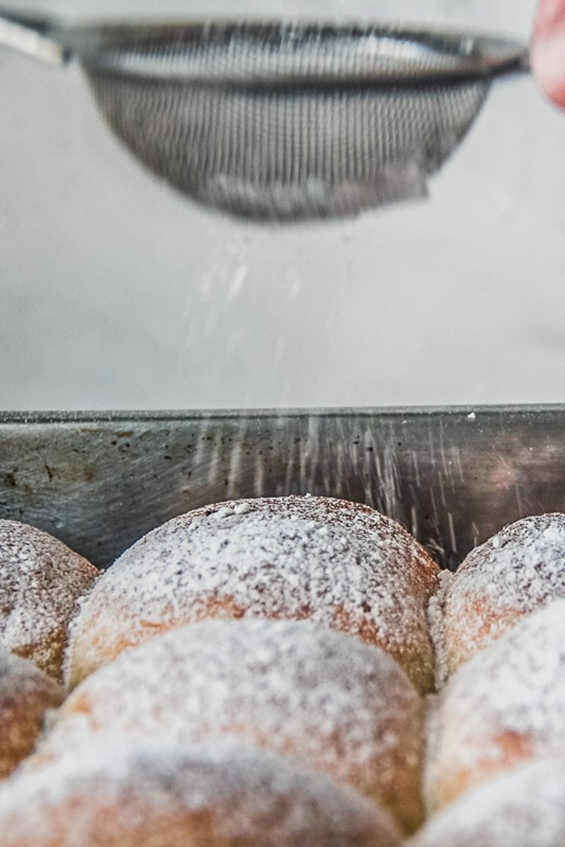 Powdered sugar is sprinkled over filled rolls in a stainless steel baking tin.