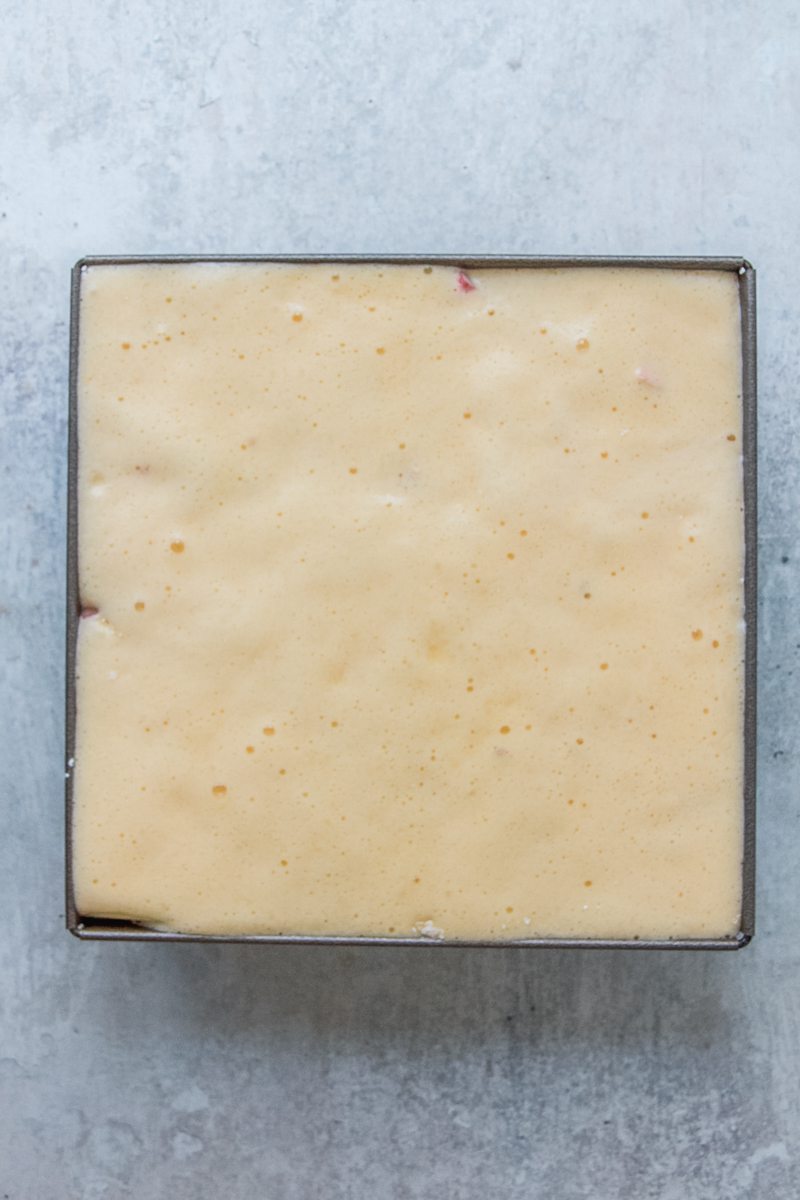 Whipped sponge cake sits on top of rhubarb in a square metal cake tin on a gray surface.