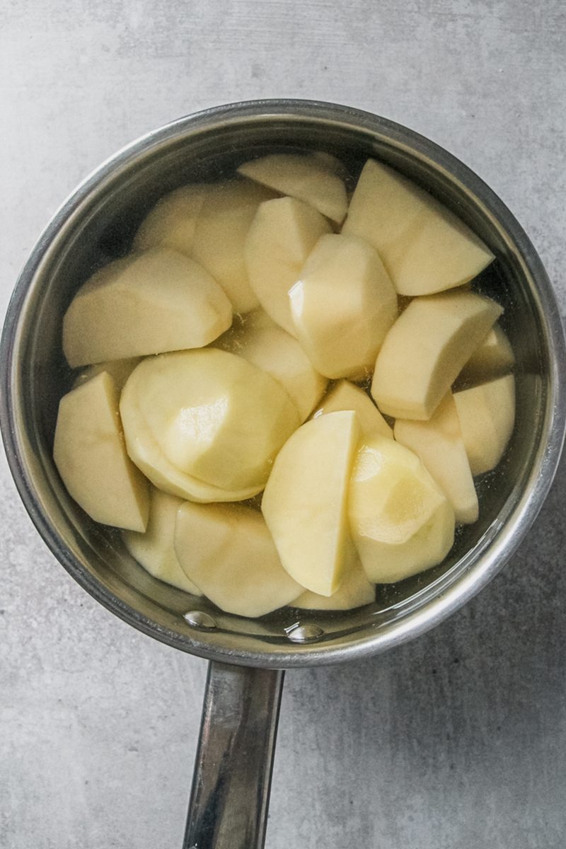 Roughly chopped potatoes sit in a stainless steel saucepan covered with water on a gray surface.