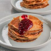 A stack of golden Icelandic pancakes sit on a rimmed ceramic plate with a cube of butter and heavy dollop of jam rolling off the stack on a gray surface.