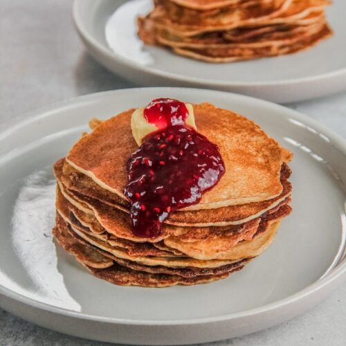 A stack of golden Icelandic pancakes sit on a rimmed ceramic plate with a cube of butter and heavy dollop of jam rolling off the stack on a gray surface.