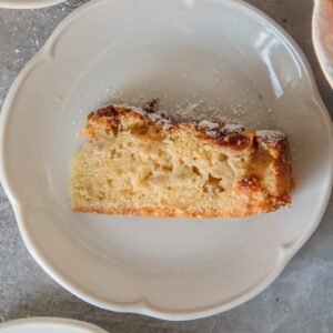 A slice of Irish apple cake sits served up on a lightly scalloped white plate on a gray surface.