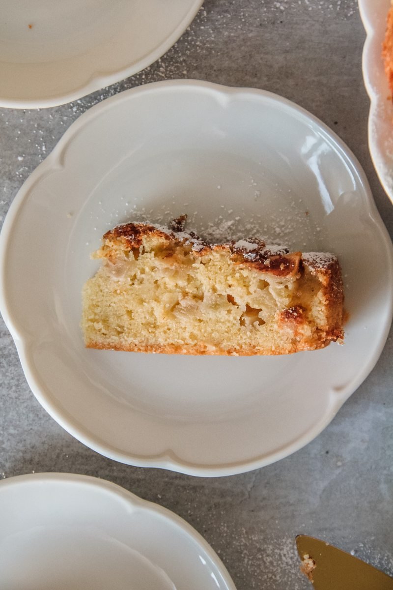 A slice of Irish apple cake sits served up on a lightly scalloped white plate on a gray surface.