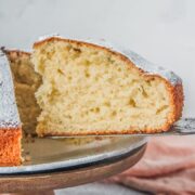 A slice of Irish Tea Cake sits on a cake server from a fluted white ceramic plate on a wooden cake stand on a gray surface.