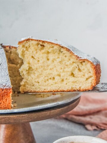 A slice of Irish Tea Cake sits on a cake server from a fluted white ceramic plate on a wooden cake stand on a gray surface.