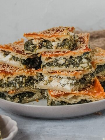 Spanakopita or spinach and feta borek slices sit stacked on a rimmed gray ceramic plate on a gray surface.