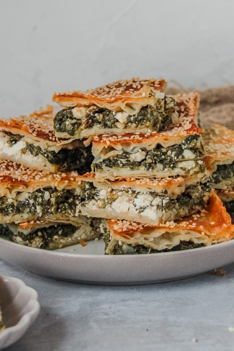 Spanakopita or spinach and feta borek slices sit stacked on a rimmed gray ceramic plate on a gray surface.