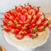 Halved strawberries sit on top of a three layer sponge cake decorated with whipped cream on a white fluted cake stand on a gray surface.