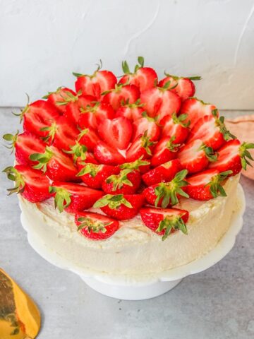 Halved strawberries sit on top of a three layer sponge cake decorated with whipped cream on a white fluted cake stand on a gray surface.