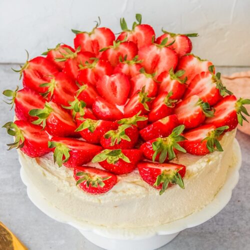 Halved strawberries sit on top of a three layer sponge cake decorated with whipped cream on a white fluted cake stand on a gray surface.