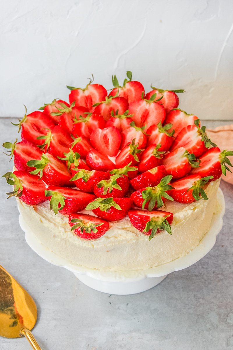 Halved strawberries sit on top of a three layer sponge cake decorated with whipped cream on a white fluted cake stand on a gray surface.