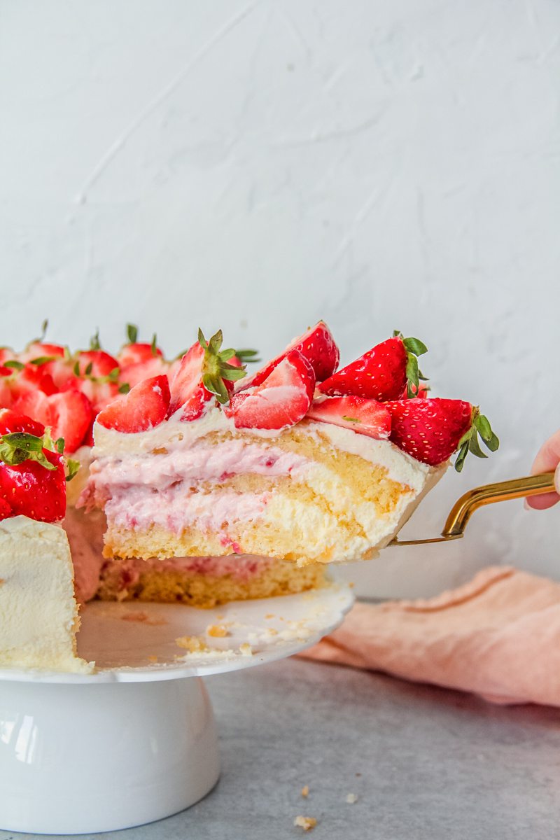 A slice of Jordgubbstarta - Swedish Strawberry Cake sits on a cake server beside the remaining cake with the strawberry and cream center visible.